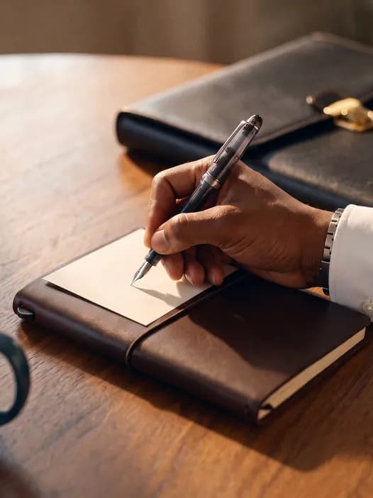 Mãos anotando em um caderno ao lado de uma pasta de couro